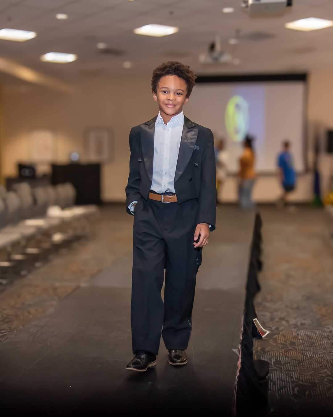 A young boy in a black tuxedo with tails walks a runway past rows of empty chairs at a fashion show.