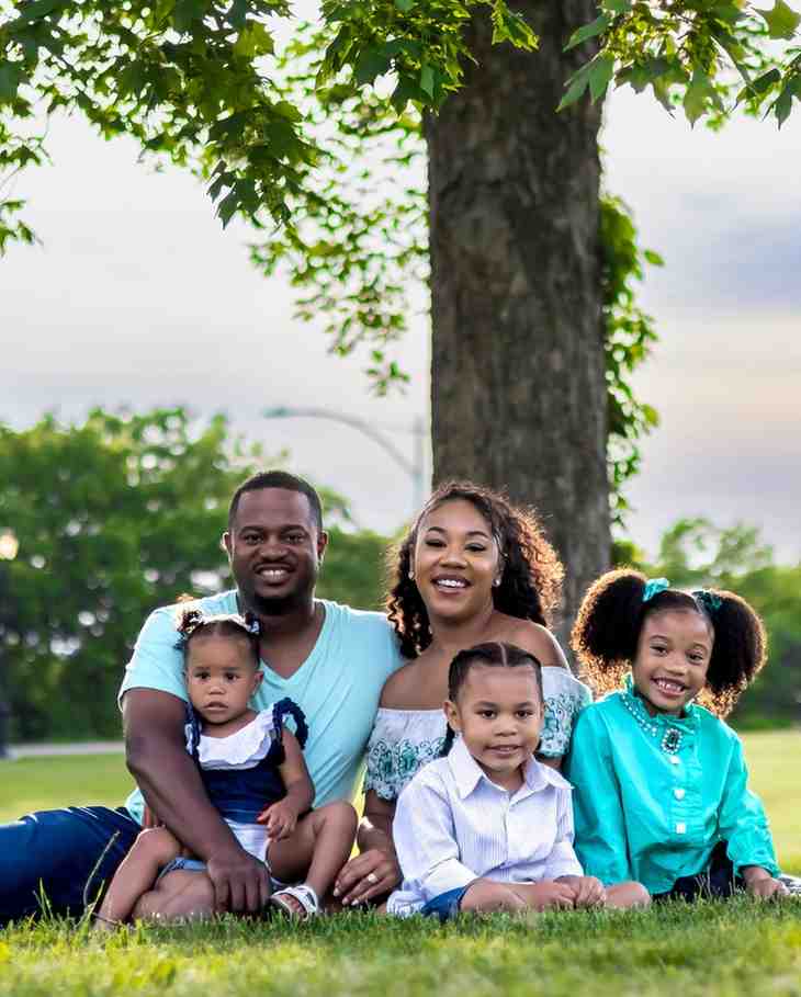 A family of five, two parents and three young children, sit together on grass under a large tree, smiling at the camera.