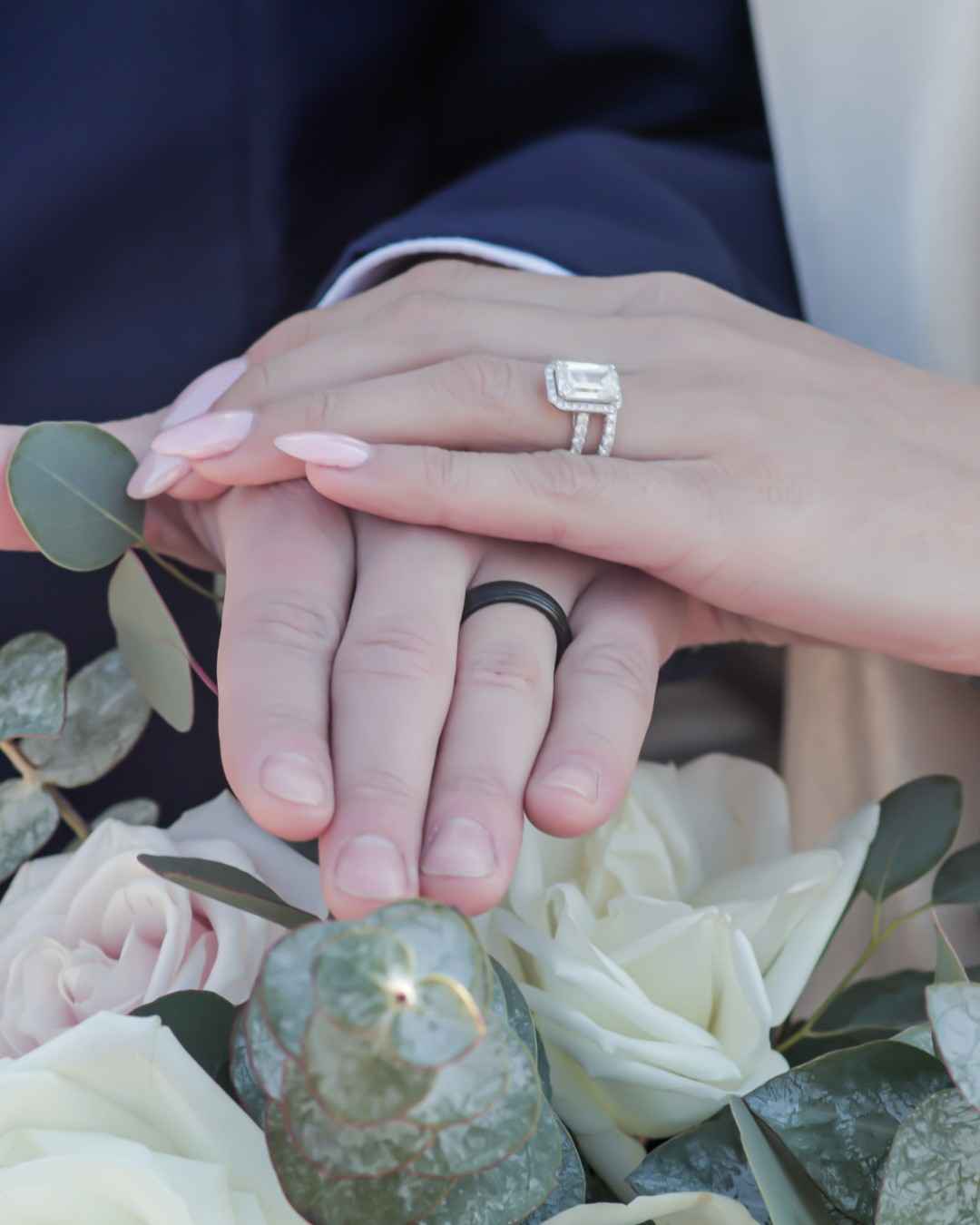 Close-up of two clasped hands with a diamond wedding ring set and a black wedding band, resting on white roses and eucalyptus.